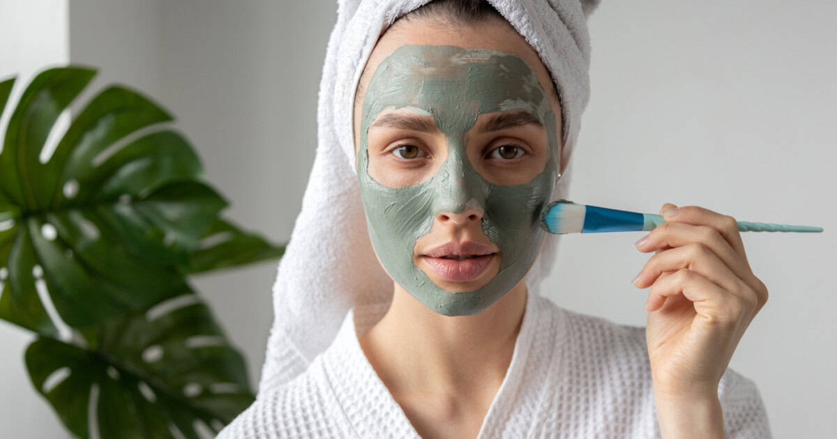 Jeune femme portant une serviette sur la tête et un peignoir mettant un masque d'argile sur le visage avec une brosse regardant la caméra.