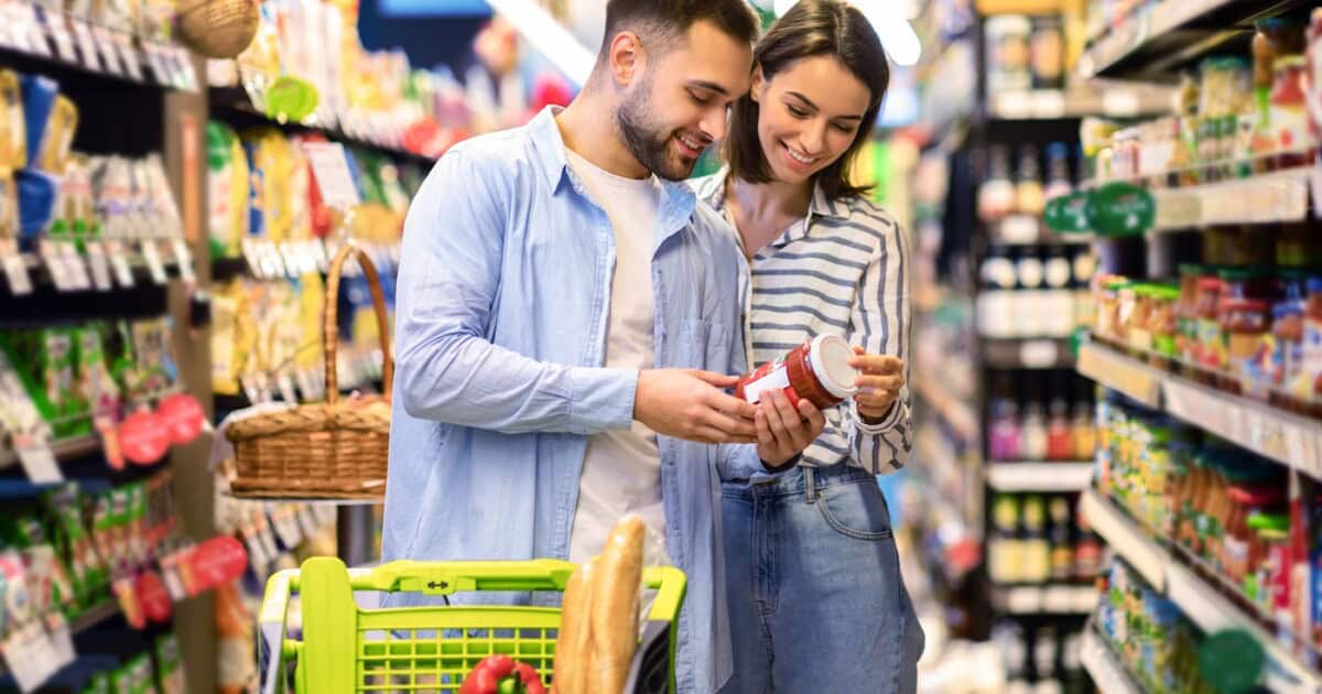 Heureux couple achetant de la nourriture dans le supermarché, en choisissant des produits debout avec un chariot de chariot le long des allées et des étagères complètes achetant ensemble des produits d'alimentation.
