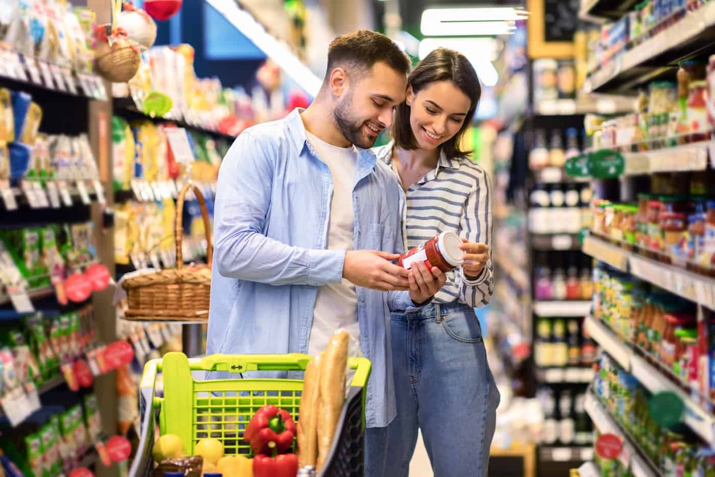 Heureux couple achetant de la nourriture dans le supermarché, en choisissant des produits debout avec un chariot de chariot le long des allées et des étagères complètes achetant ensemble des produits d'alimentation.