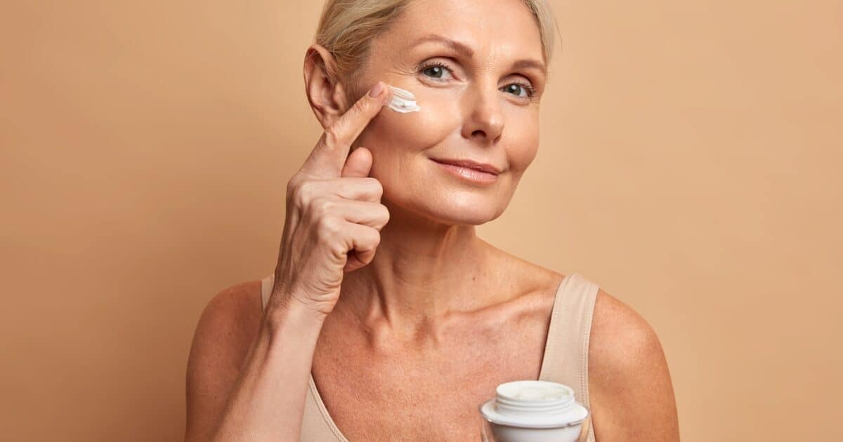 Gros plan d'une belle femme d'âge moyen applique une crème anti-âge sur le visage et subit des soins de beauté qui se soucient des poses de peau sur fond beige. Modèle féminin ridé avec produit cosmétique