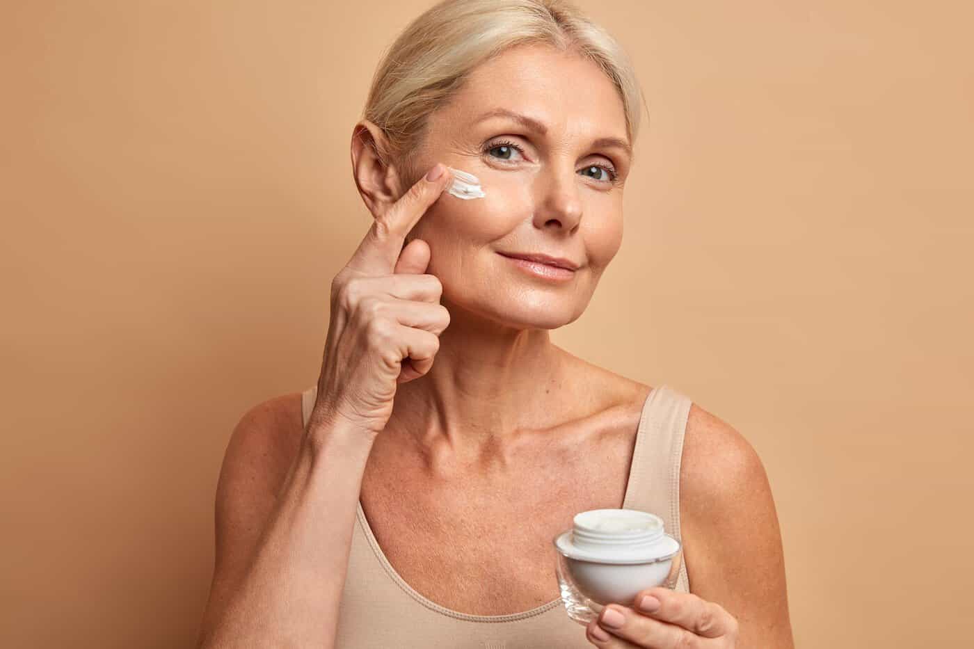 Gros plan d'une belle femme d'âge moyen applique une crème anti-âge sur le visage et subit des soins de beauté qui se soucient des poses de peau sur fond beige. Modèle féminin ridé avec produit cosmétique