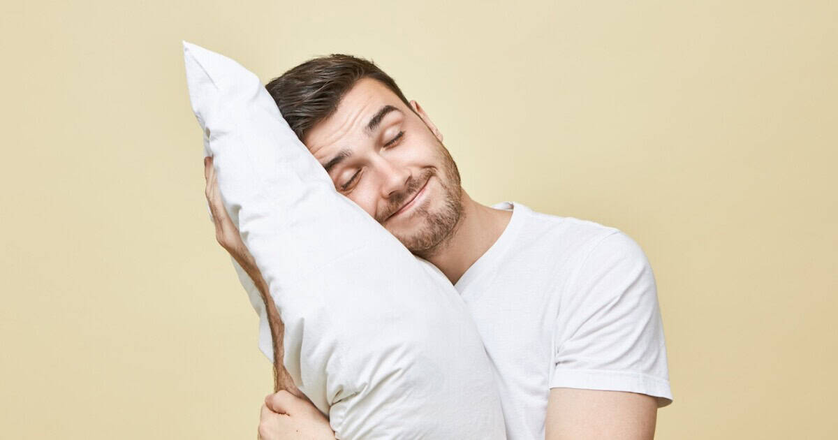 Homme heureux câlinant un oreiller blanc reconditionné, les yeux fermés, illustrant le confort et la qualité du sommeil sur fond beige