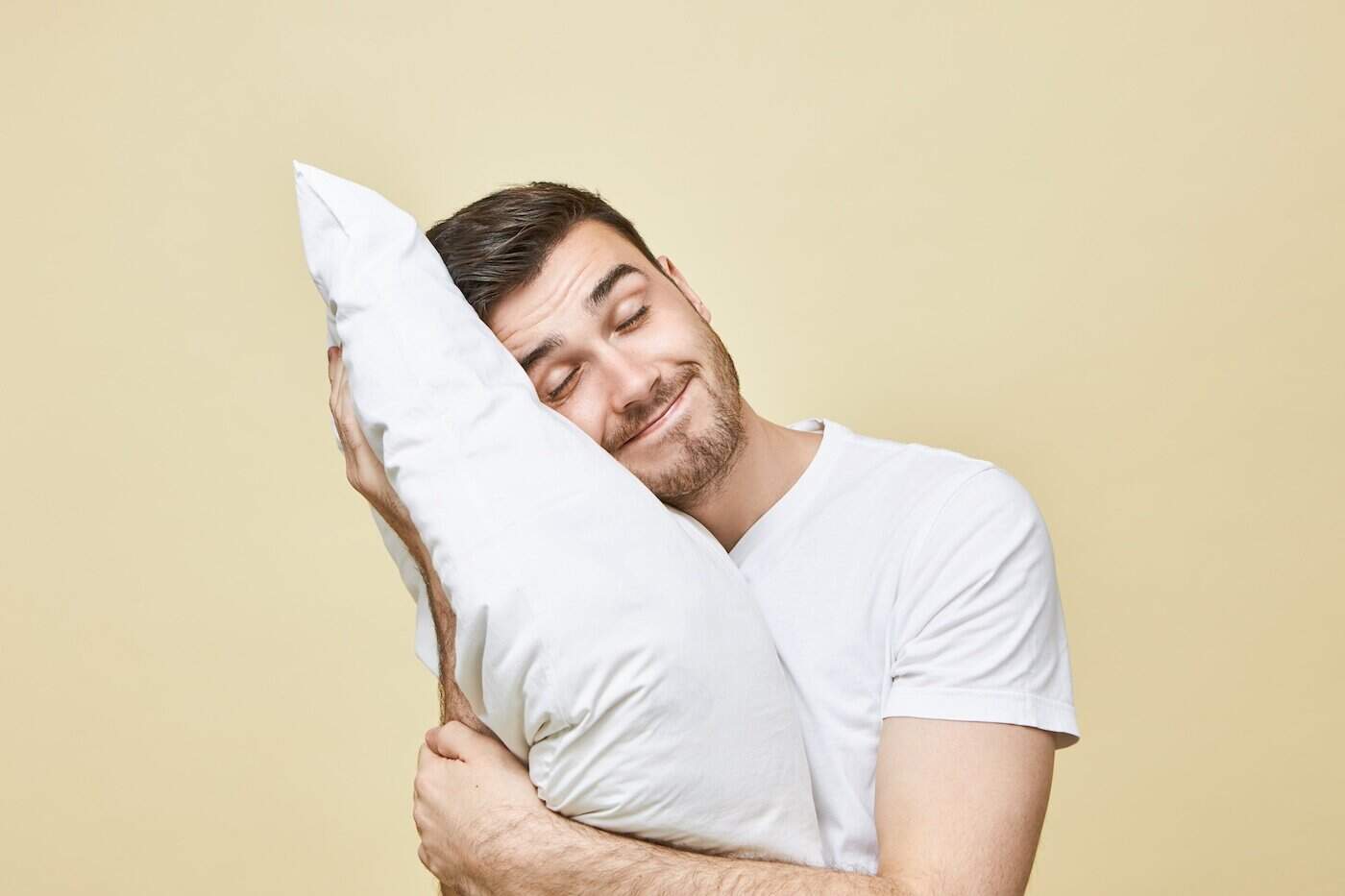 Homme heureux câlinant un oreiller blanc reconditionné, les yeux fermés, illustrant le confort et la qualité du sommeil sur fond beige