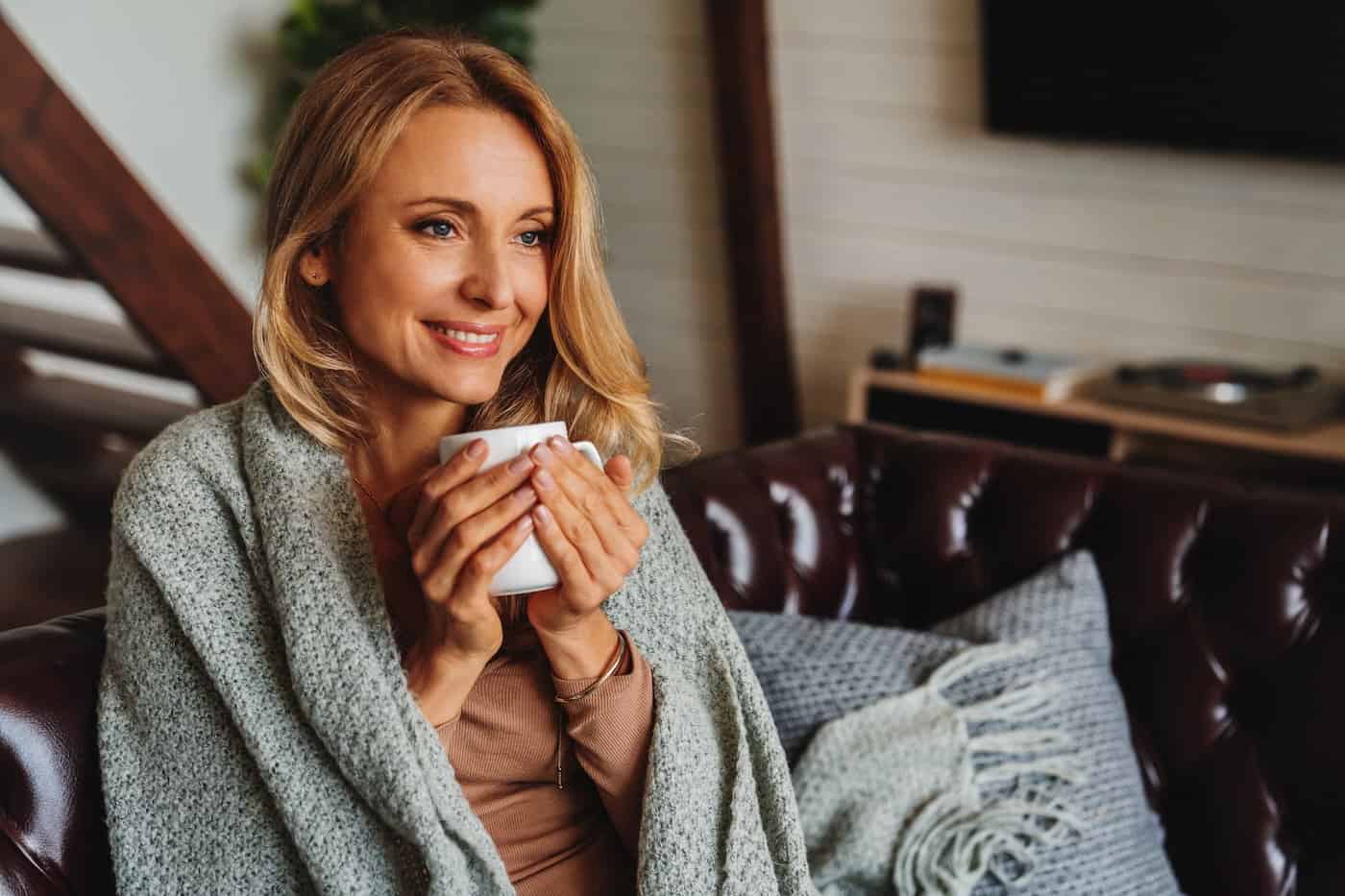 Gros plan d'une femme d'âge moyen rêvant assise dans le salon avec une tasse de café ou de thé sous une couverture